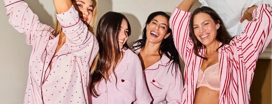 A Group of 4 women of varying ages wearing pink and red pyjamas with hearts and a fluffy robe standing on a bed like a PJ party