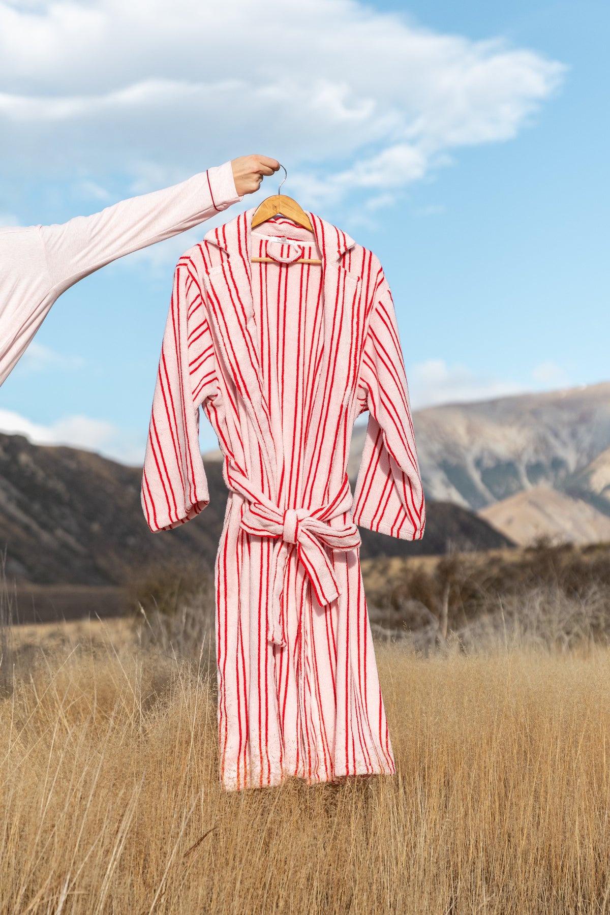 A red and white vertically striped bathrobe with a tie belt hangs from a wooden hanger held by a person's arm, set against a blurred outdoor background of a grassy field and mountains.