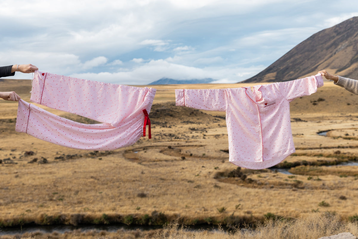 A pink patterned kimono robe and matching skirt are hanging on a string outdoors over a dry grassy landscape with mountains in the background. Hands are visible holding the garment on each side.